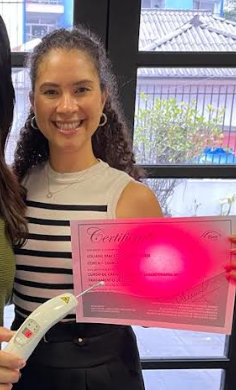 Imagem da autora, cor de pele branco, com cabelos cacheados, blusa listrada, em pé, segurando o certificado do curso de laser. 