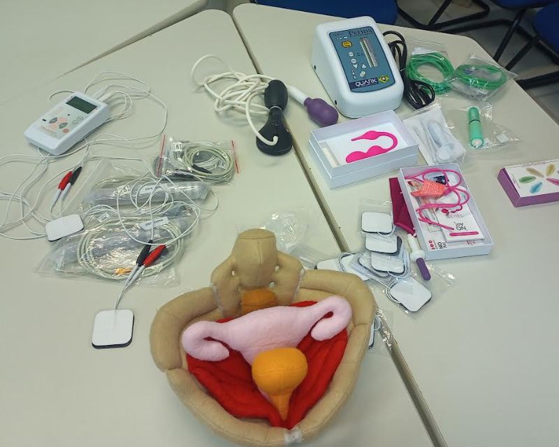 Uma mesa, com vários equipamentos para a reabilitação do assoalho pelve. Possui uma pelve de tecido, um equipamento de eletroestimulação, cones vaginais, eletrodos e aparelho de biofeedback.