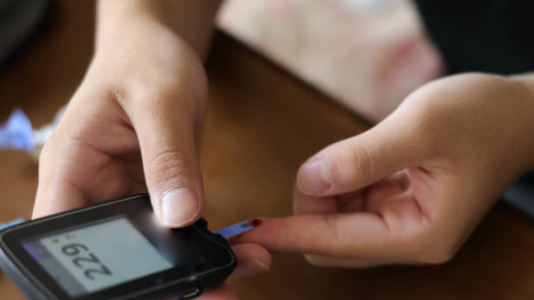 Fotografia das mãos de um indivíduo realizando o controle da glicemia com o glicosimetro, que registra 229 mg/dL no visor.
