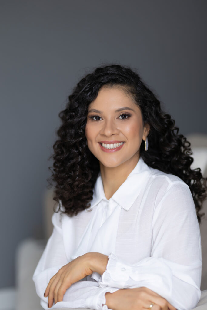Imagem de uma mulher branca, sentada em um sofá, com cabelos cacheados, brinco cor branco, blusa cor branca, está sorrindo.